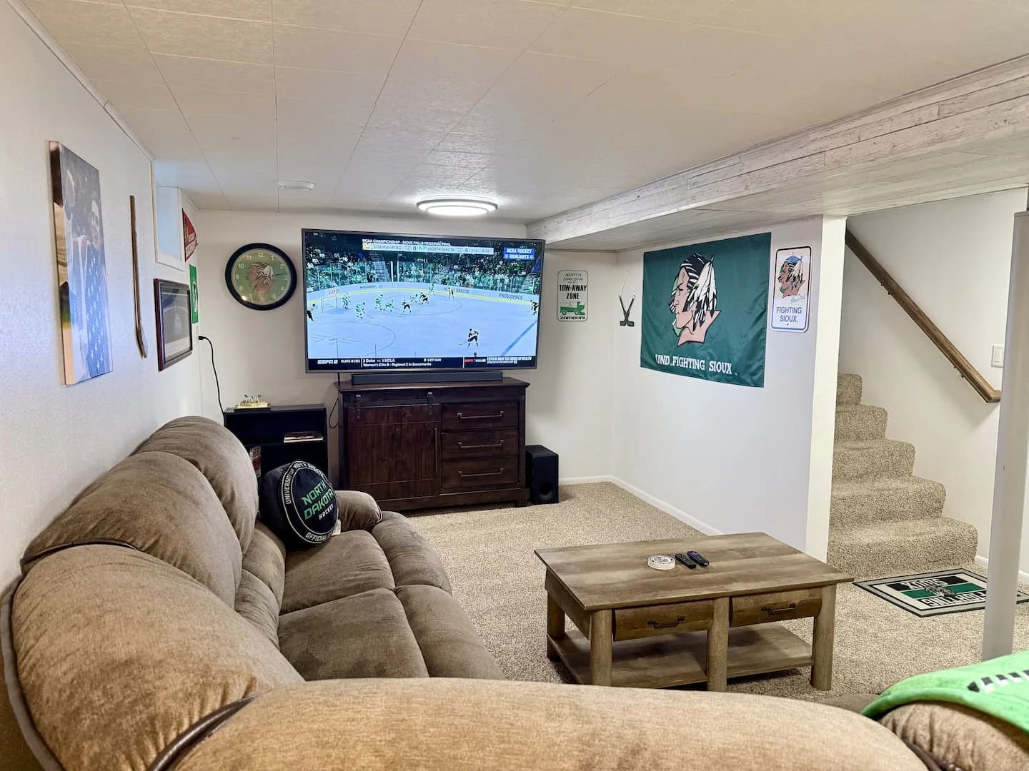 Downstairs family room with a brown sectional, coffee table, large TV and soundbar, hockey decor, and stairs on the right