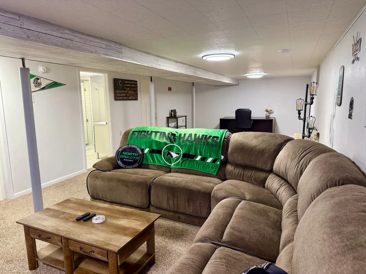 Brown sectional with a green Fighting Hawks blanket and puck-shaped cushion, with a desk and office chair behind it
