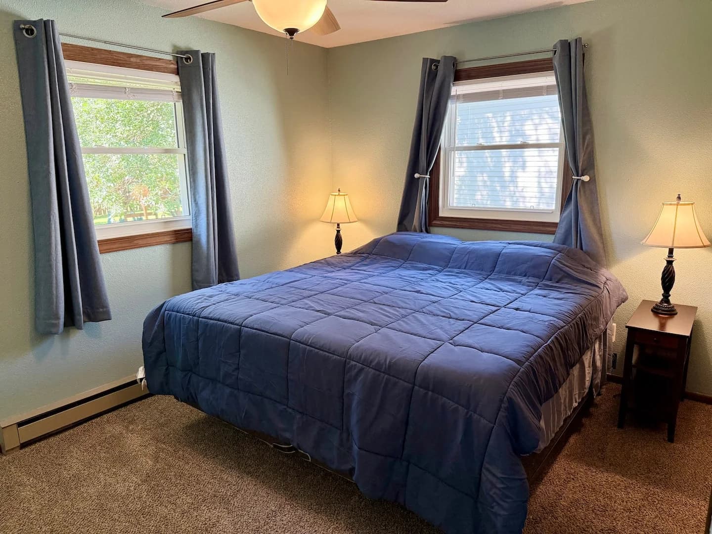 King bedroom with blue bedding, bedside lamps, and two windows with dark curtains