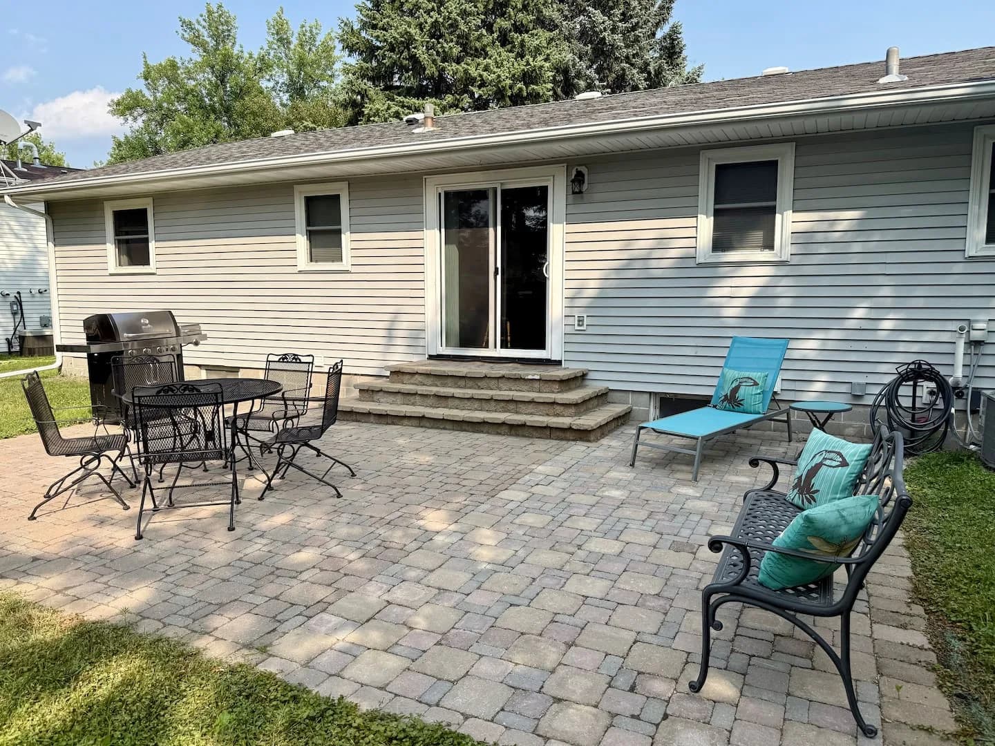 Backyard paver patio with a grill, dining table and chairs, bench, lounge chair, and steps to the sliding door